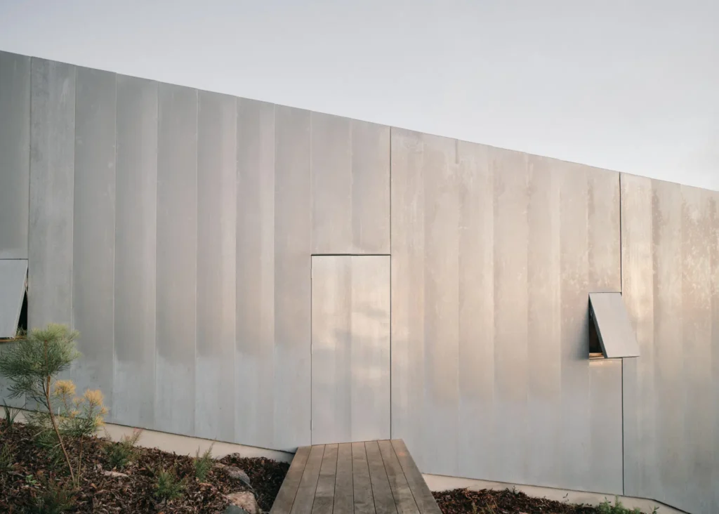 Curved zinc-clad building facade with a flush door, small shuttered window, and wooden walkway amid planted landscaping