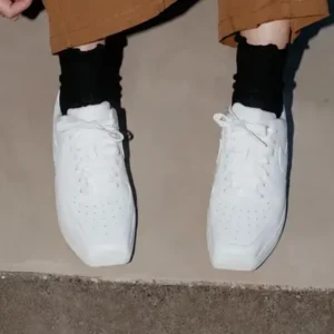 White low-top shoe with perforated toes, black socks, and brown cropped pants against a concrete floor