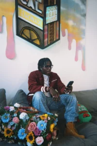Nino Paid sits on a couch holding a phone, wearing a burgundy jacket, sparkly blue jeans and brown boots, with flowers