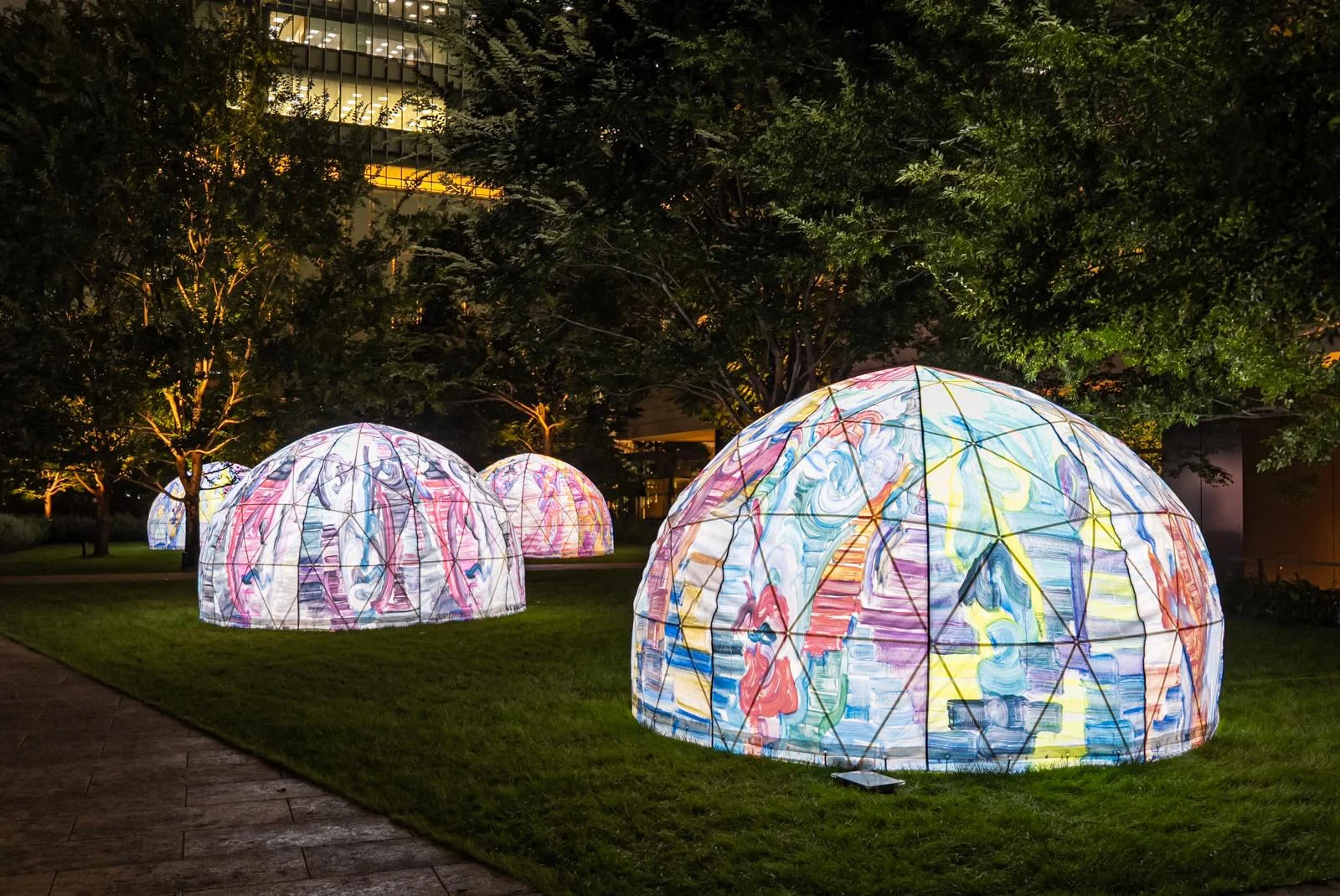 Illuminated dome-shaped sculptures covered in colorful abstract drawings sit on a lawn at night, with trees and office