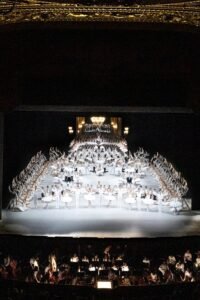 Large ensemble of ballet dancers in white forms a pyramid on a lit stage above a visible orchestra pit