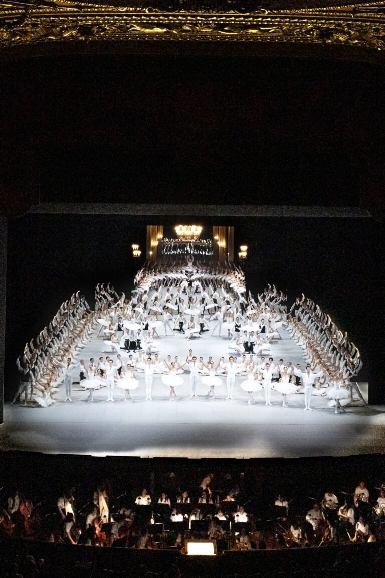 Large ensemble of ballet dancers in white forms a pyramid on a lit stage above a visible orchestra pit