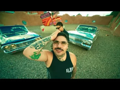 That Mexican OT in a black tank top poses with his hand on his head between two vintage cars in a gravel lot