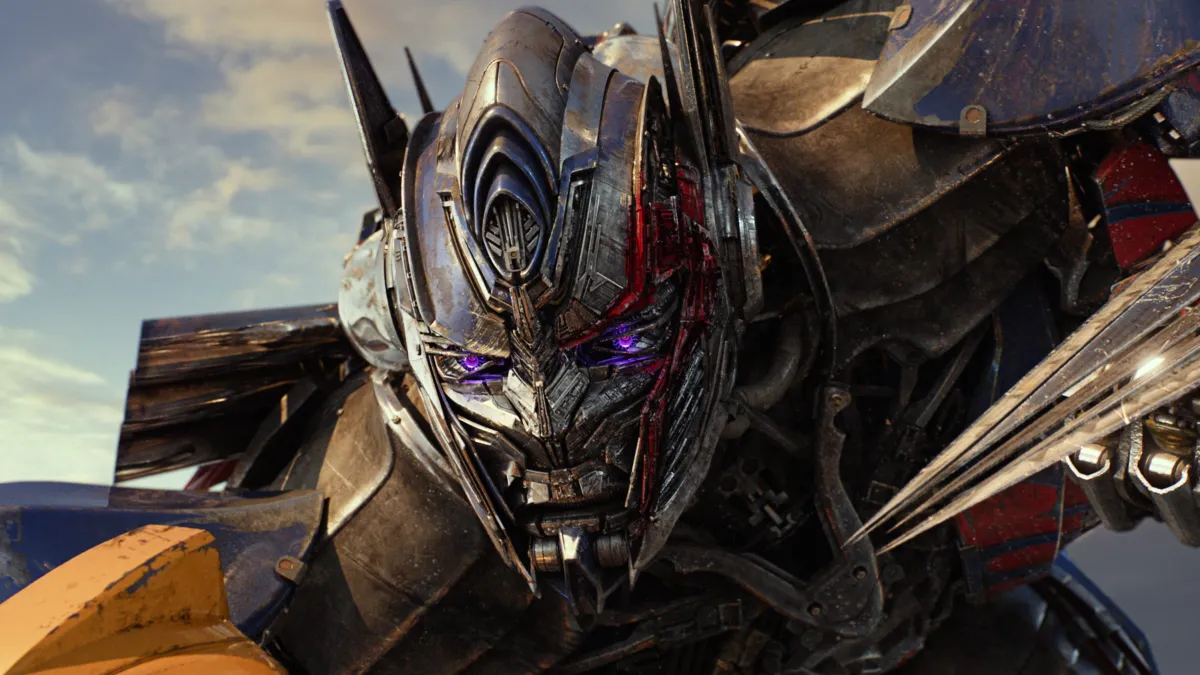 Close-up of a battle-worn metallic robot with glowing purple eyes, angular armor, and red and blue panels against the sky