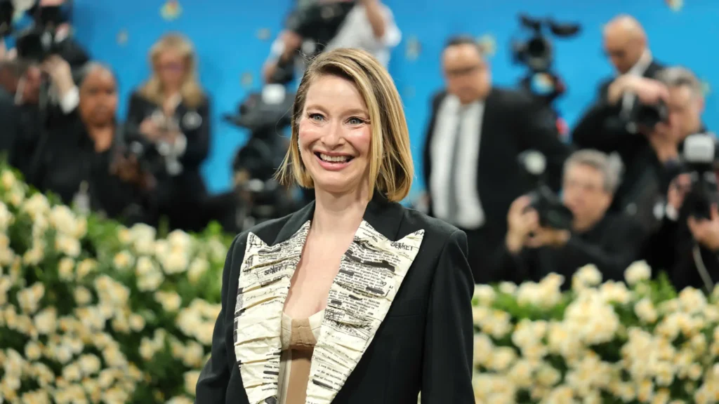 Hildy Kuryk in a black blazer with cream newspaper-print lapels, posed before photographers and white flowers on a blue