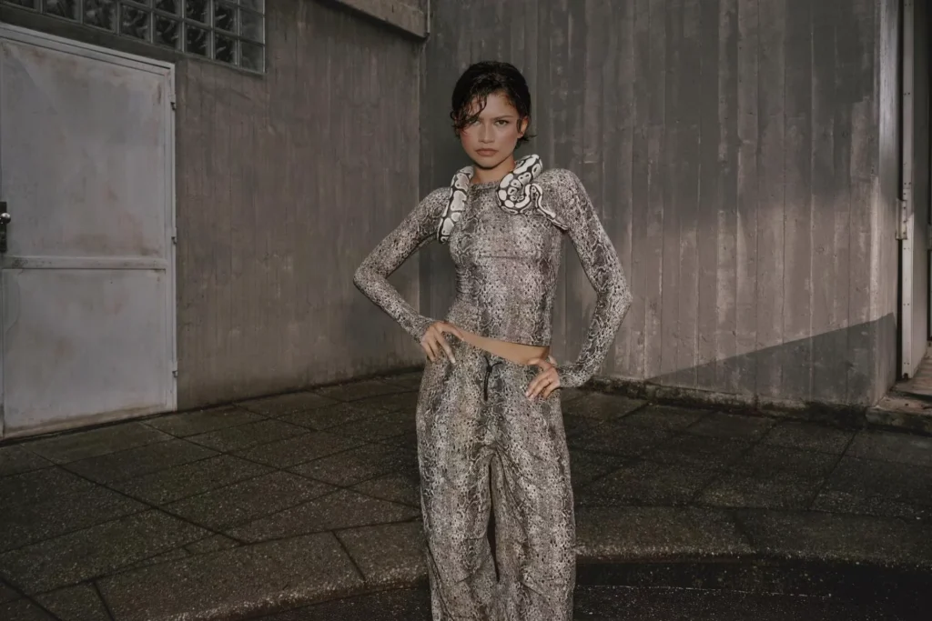 Zendya poses with hands on hips in a gray snakeskin-print top and wide-leg pants beside a weathered concrete wall