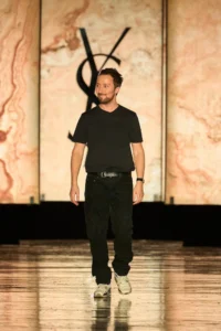 Bearded man in a black Tee and pants walks on a reflective runway before abstract peach-toned panels