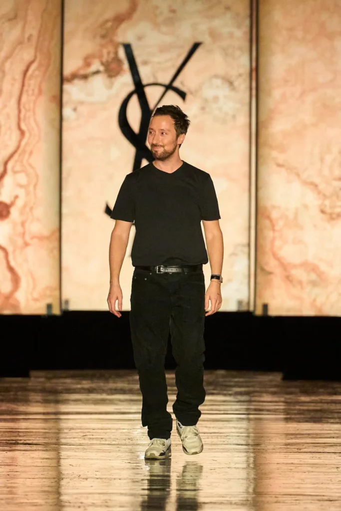 Bearded man in a black Tee and pants walks on a reflective runway before abstract peach-toned panels