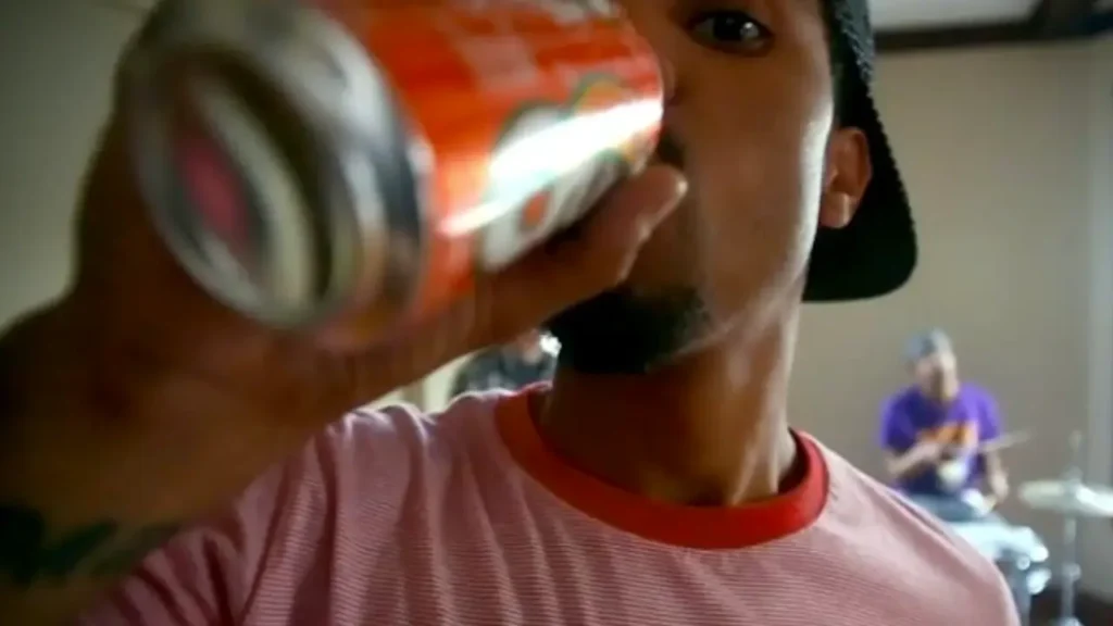 Vic Mensa in a pink shirt drinks from an orange soda bottle, with a guitarist blurred in the background