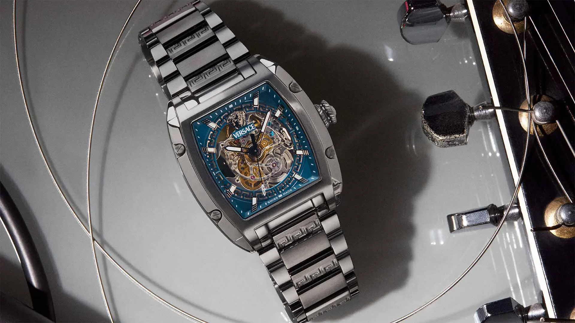 Versace skeleton watch with a blue dial and dark metal bracelet displayed on a reflective stand beside jewelry hardware