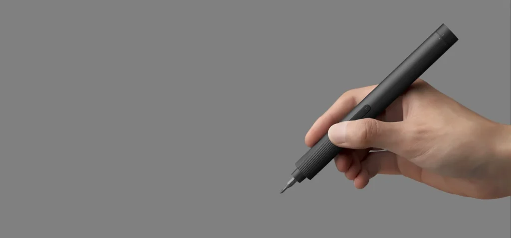 Hand holding a black precision cordless screwdriver with a ridged grip and exposed bit against a gray background